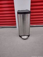 Full front view of cylindrical stainless steel trash bin with pedal at base, black lid and base visible, no damage seen.