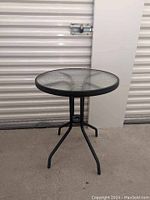 Full view of the small round black metal frame table with textured glass top on concrete floor.