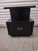 Front view of a small black nightstand showing drawer with metal handle and elevated top surface.