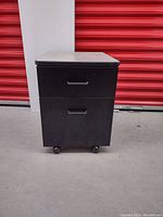 Front view of black nightstand showing two drawers with black handles and caster wheels, set on concrete floor with red storage door in background.