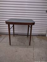 Front view of the vintage folding table showing wooden legs and blue-gray tabletop against a metal door background.