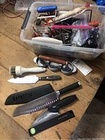 Photo showing multiple kitchen knives, including some in sheaths, a white plastic masher, patty maker, and a storage container with various utensils inside.
