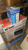 Boxed Mr. Heater and Dyna-Glo tank top propane heaters stacked showing full size and front of packages.