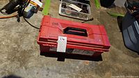 Closed red plastic toolbox on concrete floor, shows external condition and handle.