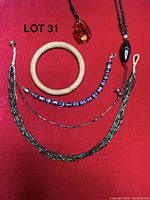Photo showing multi-strand green and black glass bead necklace, olive wood bangle bracelet, amber pendant on cord, blue glass bead bracelet, and thin bead strand bracelet on red background.