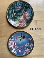 Top view of two hummingbird collector plates with colorful floral and hummingbird designs and gold trim on rim, shown on wooden floor