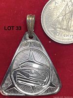 Triangular silver pendant with eagle design next to a Canadian 10 cent coin for scale, engraved in traditional indigenous style.