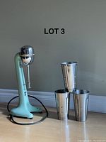 Full shot of milkshake maker base and three stainless steel cups upright on floor against wall