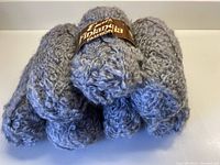 Seven balls of soft nubbly wool yarn piled with one ball showing a brown label with brand info.