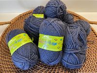 Nine balls of soft blue/grey yarn in a basket, some tightly wound in skeins with green labels showing the Shepherd Colour 4 Me branding.