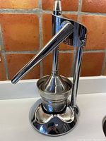 Front view of the chrome manual juicer showing lever handle, cone press, and cup inside base.