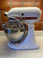 Front left side view of the white KitchenAid stand mixer with stainless steel bowl and wire whisk attached.