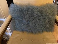 Rectangular grey sheepskin pillow with long curly wool resting on beige cushioned chair, showing full size and overall condition.