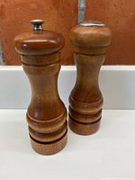 Both pepper grinder and salt shaker shown side by side on white surface with brick background displaying overall condition and finish.