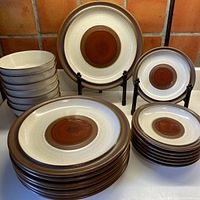 Photo showing stacks of 8 dinner plates, 8 luncheon plates, and 8 cereal bowls with characteristic cream speckled background, chocolate rims, and cinnamon rust center circles.