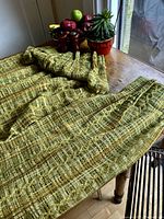 Full view of a muted lime green woven drapery panel with amber and cinnamon threads laid out on a table near a window and some decorative items.