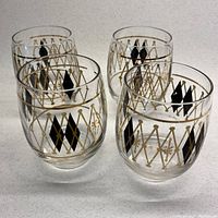 Four clear glass bar glasses with black diamond patterns and gold crisscross lattice and fleur-de-lis detailing on a white background.