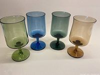 Four colored stemmed aperitif glasses in green, blue, teal, and amber shown together in one image.