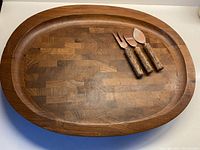 Full view of oval wooden cutting board with raised rim and three wooden handle utensils placed on top.