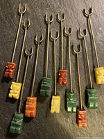 Full set of 11 hors d'oeuvre forks laid out on a dark surface showing metal fork shafts with Bakelite handles in three colors: green, orange, yellow, each with abstract carved faces.