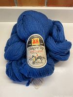 Three skeins of vivid blue-grey 100% wool Fingertip Shetland yarn laid together with visible label showing Merkels & Kienlin brand and equestrian image