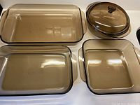 Four PYREX smokey brown glass ovenware pieces arranged on a flat surface: large rectangular dish, smaller rectangular dish, square dish, and round lidded casserole dish.