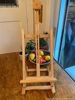 Full front view of folding wooden artists table easel on wooden table with some plants and fruit bowl in background