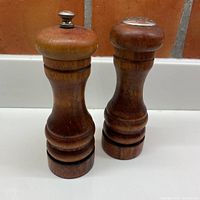 Full view of both Baribocraft salt and pepper grinders side by side on a white surface in front of a tile backsplash.