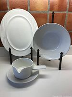Full view of lot: oval platter, large bowl, gravy boat with saucer all featuring white ceramic and indented linear lines.