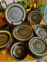 Overall view of the entire collection showing various plates, bowls, and cups with saucers, arranged on a wooden surface with plants in background.