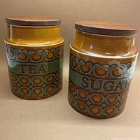 Front view of the tea and sugar ceramic cannisters with wooden lids, showing the decorative pattern and lettering.
