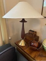 Full view of lamp with teak wood body, brass base, and off-white fabric shade sitting on a wooden table next to small wooden boxes and a carved bowl.