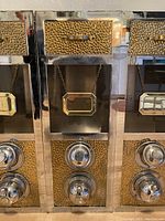 Front view of the vintage dispenser showing the hammered brass drawer front with chrome knobs and a hanging brass-framed name tag on chain.