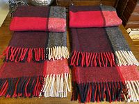 Two folded red, black, cream, and dark gray check wool blankets with fringed ends laid side by side showing matching pattern and fringe details.