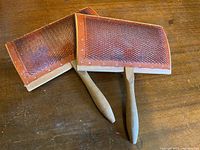 Two wooden carding combs with curved backs and handles placed on wooden surface, showing front side with metal bristles.