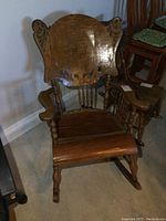 Front view of antique oak rocking chair showing carved high back with some veneer peeling on seat and missing spindles in backrest.