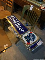 Photo showing three plastic beer signs placed on chairs: large Pabst Blue Ribbon rectangular sign, Schlitz Malt Liquor bull sign, and round Pabst Blue Ribbon sign.