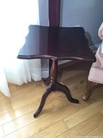 Photo showing the full small Bombay tilt-top side table with its scalloped square top and tripod pedestal base, placed on a wooden floor next to a curtain and upholstered chair.