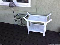 Photo shows a white painted wood cart with peeling paint and two slatted shelves on caster wheels next to a black metal table with glass top and a tall black metal candle stand.