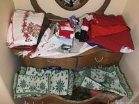 Christmas-themed table cloths, napkins, assorted ornaments including Santa figurines, placemats, and holiday decorations placed on wooden furniture.