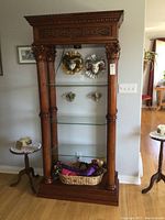 Full front view of tall wooden display shelf with carved columns and glass shelves. Top lighting visible.