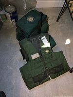 Full view of the green Sportsman flotation vest with zippered pockets and attached flotation material inside. The vest is lying on a concrete floor with the green carry bag behind it.