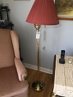 Full view of one brass floor lamp with a red pleated shade next to a chair and table.