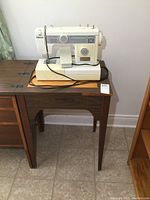 Full view of white-beige Omega sewing machine on wooden sewing stand against white wall, with foot pedal visible.
