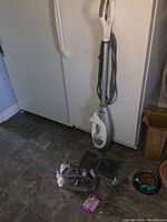 Full view of Shark Lift-Away vacuum upright unit with cord wrapped around handle, standing in front of white refrigerator on tiled floor. Accessories including hose, cleaning tool and small packaged item placed adjacent on floor.