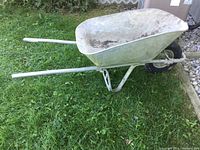 Side view of an older aluminum wheelbarrow sitting on grass, showing large aluminum tray and handles.