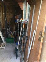 Photo showing a variety of long-handled yard tools including rakes, shovels, post hole diggers, and a broom leaning inside a shed.