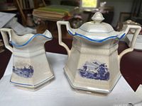 Photo showing creamer and sugar bowl side by side, displaying shape and blue Dutch scenic artwork with crackle glaze and blue rim accents.