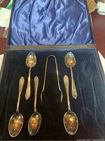 Full view of the silverware set in the open box showing 4 spoons and 1 pair of tongs, blue satin lining on the lid, and black velvet base.