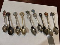 Photo showing nine collectible Canadian souvenir spoons arranged in a row, each with a decorated crest on the handle top representing different Canadian cities.